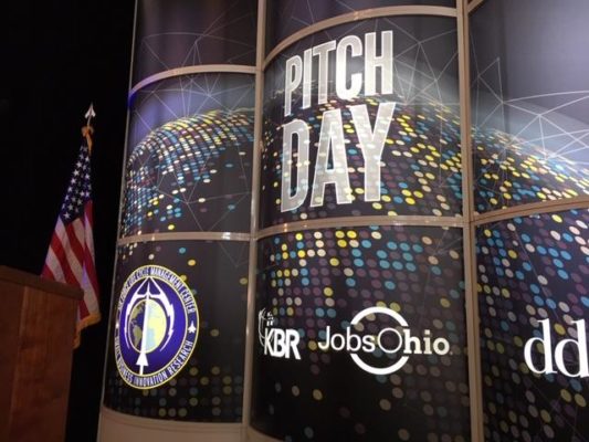 A stage setting features a large cylindrical backdrop prominently displaying "PITCH DAY" in bold white letters against a design of interconnected multicolored dots, resembling a digital globe. Below, various logos are visible, including one of the Department of Defense with a blue and gold emblem. To the left stands the American flag on a pole, its stars and stripes visible against a darker ambiance. Other logos, such as "KBR," "JobsOhio," and "dd" are also present on the backdrop. The entire setting conveys a formal event atmosphere related to defense and innovation pitches.