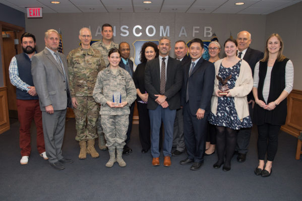 A diverse group of twelve individuals stands side by side in a well-lit room with a blue background, displaying an emblem with the text "HANSCOM AFB". Two individuals in the center wear U.S. military uniforms; a female soldier in the forefront holds a clear trophy, and a taller male soldier in camouflage stands beside her. To their left and right are civilians in business attire, some with awards in hand. They exhibit a mix of pride and formality. The overall ambiance suggests a recognition or award ceremony within a Department of Defense facility.