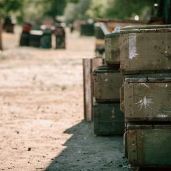 The image depicts a stack of green military ammunition boxes in a field. The boxes are rectangular, weathered, and worn, with metal clasps on the front. In the blurred background, other boxes and equipment can be seen. The image is taken during the day under natural lighting.