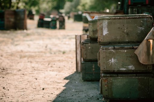 The image depicts a stack of green military ammunition boxes in a field. The boxes are rectangular, weathered, and worn, with metal clasps on the front. In the blurred background, other boxes and equipment can be seen. The image is taken during the day under natural lighting.