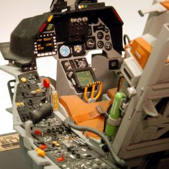 The image displays the cockpit of a United States military aircraft. The cockpit is filled with various control panels, buttons, and switches. The dashboard is black with orange and yellow accents. There is a gray seat with an orange cushion. The exterior of the cockpit is white with gray accents. The background is blurred and out of focus, emphasizing the detail of the cockpit.