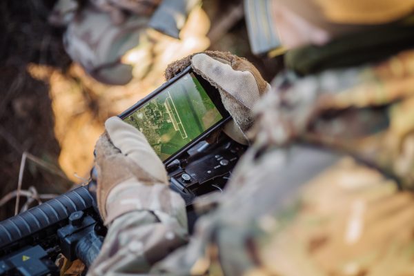 A close-up view of a soldier's gloved hands holding a compact electronic device displaying a green-tinted tactical interface with crosshairs. The soldier wears camouflage attire, and the environment suggests an outdoor setting with some foliage visible. The scene emphasizes careful operation and focus on the device's screen.
