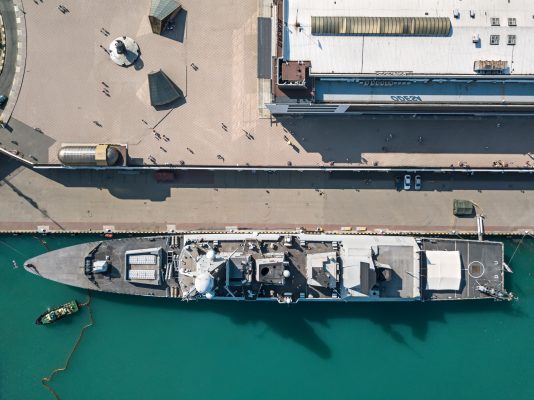 A bird's-eye view of a naval vessel docked alongside a large port. The ship, with a streamlined design, features multiple compartments and equipment on deck. Directly adjacent to the ship, the port has a spacious paved area with a few scattered structures and people walking about. Parallel to the ship, the dockside has large industrial buildings with varying rooftops. The surrounding water is a clear turquoise, and a smaller boat is seen near the ship, possibly aiding in docking procedures. The entire scene depicts a calm day at a military or naval port.