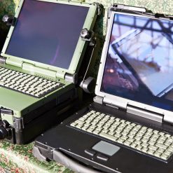 Close-up view of two military-grade rugged laptops placed side by side. Both laptops have reinforced hinges and sturdy chassis, with one being a dark green color and the other in matte black. They feature heavy-duty latches, tactile keyboards, and thick bezels around their screens. One laptop has its screen displaying an unclear image, possibly of a tent structure, while the other has a darkened screen. The laptops rest on a fabric with a green and brown camouflage pattern, hinting at their use in field operations. The overall design prioritizes durability and resilience for demanding environments.