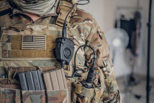 A close-up view of a soldier's torso, highlighting the details of their camouflage tactical vest. Prominently displayed is a weathered U.S. flag patch, stitched in subdued colors, signifying their affiliation with the United States military. Directly beneath it, there are two ammunition magazines, one in dark gray and another in a light tan, securely nestled in their respective pouches. To the right, a black, circular military communication device, possibly a PTT (Push-To-Talk) unit, is tethered to the vest. Attached to it is a spiraled black cord leading to a clip-on attachment, suggesting a hands-free communication system. The top portion of the image shows a hint of a beige scarf wrapped around the soldier's neck. In the blurred background, a room with a standing fan and other indistinct objects can be observed, suggesting an indoor setting, possibly a base or a briefing room.
