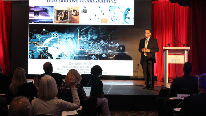 A man stands to the right of a large presentation screen, addressing an audience. The screen displays various graphics related to additive manufacturing, including vehicle parts and digital interfaces. The title "DOD Additive Manufacturing" is prominent at the top. Below, text introduces "Dr. Alan Pentz" with a title indicating his position. The audience, with some members visible in the foreground, attentively watches the presenter who stands behind a podium with a draped cloth. The ambiance suggests a professional conference setting.