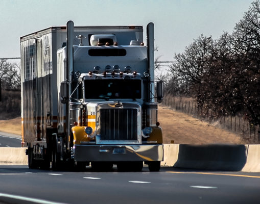 A large semi-truck travels on a sunlit highway. The truck has a shining chrome front, featuring a prominent grille and multiple overhead lights. It's attached to a sizable, rectangular cargo trailer that appears slightly weathered. To the truck's right, a fence runs alongside a barren tree line, signaling a winter or autumn season. The road, defined by clear lane markings, contrasts against the truck's imposing presence, emphasizing the vehicle's significance on the open road.