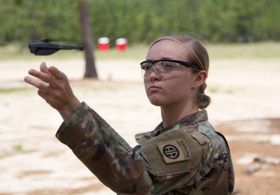 A focused individual in U.S. military camouflage attire, prominently displaying an "AIRBORNE" patch, is mid-action, appearing to gently toss a small drone. The background is a sandy ground with scattered trees and distant red objects. The person is wearing clear safety glasses and has a determined expression.