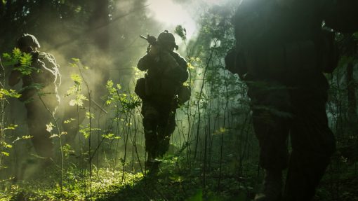 Soldiers, clad in combat gear, move stealthily through a misty forest, with sun rays piercing through dense foliage. One soldier, center-focused, kneels aiming his rifle, while others surround him in various stances.