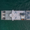 This image is an aerial view of a grey United States Navy ship at sea with a helicopter landing pad on the deck. The ship appears to be a destroyer or cruiser class vessel sailing through the water, creating a wake behind it. The ship has several gun turrets and missile launchers visible on the deck and a large radar dome on the superstructure.