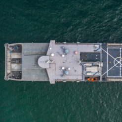 This image is an aerial view of a grey United States Navy ship at sea with a helicopter landing pad on the deck. The ship appears to be a destroyer or cruiser class vessel sailing through the water, creating a wake behind it. The ship has several gun turrets and missile launchers visible on the deck and a large radar dome on the superstructure.