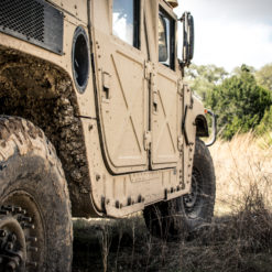 This image is a photo realistic view of a tan military vehicle parked in a field. The vehicle has large tires and appears to be covered in mud. The background consists of tall grass and trees. The vehicle is parked on a dirt road. The image is taken from a low angle, looking up at the vehicle.