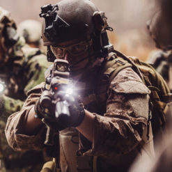 This image is a photo realistic view of United States military personnel in combat gear during a training exercise. The personnel are wearing helmets with mounted cameras and holding weapons with mounted flashlights. The personnel are wearing camouflage uniforms and body armor. The background is a wooded area with trees and foliage. The image is taken from the perspective of one of the personnel, with the focus on the person in the center of the image.