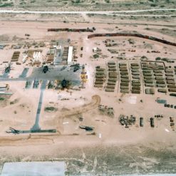This is an aerial view of a military base located in a desert environment. The base consists of several rectangular buildings arranged in a grid-like pattern, indicative of strategic planning and organization. Scattered throughout the base are various military vehicles and equipment, including tanks, trucks, and helicopters, ready for deployment. A road runs through the middle of the base, providing access to all areas. The base is enclosed by a fence for security. The surrounding terrain is primarily desert with sparse patches of grass and shrubs, typical of arid regions. This image represents the logistical and strategic aspects of military operations.