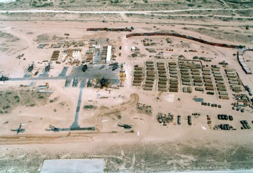 This is an aerial view of a military base located in a desert environment. The base consists of several rectangular buildings arranged in a grid-like pattern, indicative of strategic planning and organization. Scattered throughout the base are various military vehicles and equipment, including tanks, trucks, and helicopters, ready for deployment. A road runs through the middle of the base, providing access to all areas. The base is enclosed by a fence for security. The surrounding terrain is primarily desert with sparse patches of grass and shrubs, typical of arid regions. This image represents the logistical and strategic aspects of military operations.