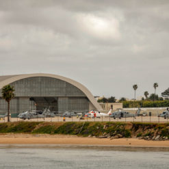 This is a photograph of a hangar housing several military helicopters. The hangar, a large structure with a curved gray roof and walls, is situated near a body of water with a sandy beach visible in the foreground. At least six helicopters of varying sizes and colors are parked on a concrete apron in front of the hangar, ready for deployment. The image, taken from across the water, captures the gloomy mood set by the cloudy sky. This scene represents the readiness and versatility of military aviation operations.