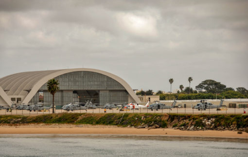 This is a photograph of a hangar housing several military helicopters. The hangar, a large structure with a curved gray roof and walls, is situated near a body of water with a sandy beach visible in the foreground. At least six helicopters of varying sizes and colors are parked on a concrete apron in front of the hangar, ready for deployment. The image, taken from across the water, captures the gloomy mood set by the cloudy sky. This scene represents the readiness and versatility of military aviation operations.