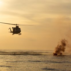 This is a photo-realistic image of a UH-1 Iroquois military helicopter, also known as a ‘Huey’, flying over the ocean. The helicopter is moving from left to right in the image. In the background on the right side, there’s a large explosion causing a plume of smoke and fire, possibly indicating a military operation or exercise. The ocean below is calm, and the sky is orange from the setting sun, creating a stark contrast with the dramatic scene. Despite the distance from which the image is taken, making the helicopter and explosion small in the frame, the scene conveys a sense of action and intensity associated with military operations.
