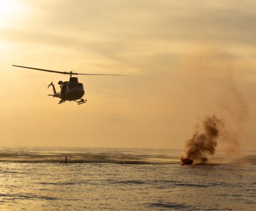 This is a photo-realistic image of a UH-1 Iroquois military helicopter, also known as a ‘Huey’, flying over the ocean. The helicopter is moving from left to right in the image. In the background on the right side, there’s a large explosion causing a plume of smoke and fire, possibly indicating a military operation or exercise. The ocean below is calm, and the sky is orange from the setting sun, creating a stark contrast with the dramatic scene. Despite the distance from which the image is taken, making the helicopter and explosion small in the frame, the scene conveys a sense of action and intensity associated with military operations.