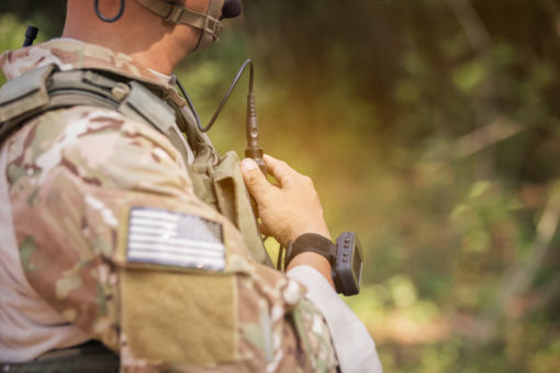 Image of a United States military personnel in camouflage uniform using a radio in a forested area. The personnel is wearing a camouflage uniform with a tan vest and a black headset. The radio is black and has an antenna. The background is blurred and consists of trees and foliage. The personnel is holding the radio with both hands and appears to be speaking into the headset.
