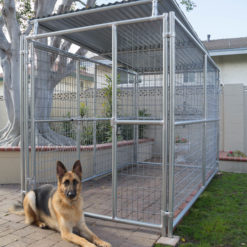A photo of a military working dog kennel with a German Shepherd dog lying in front of it. The kennel is made of metal and is rectangular in shape. The kennel has a roof and three walls, with the fourth wall being a gate. The kennel is in a backyard with a tree and a house in the background. The dog is lying on a brick patio in front of the kennel.