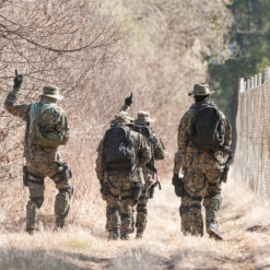 This image captures four soldiers in camouflage uniforms, walking along a path next to a wire mesh fence topped with barbed wire. They are carrying backpacks, indicating they are on a mission. The path is nestled between lush trees and bushes on one side, and the fence on the other, creating a narrow passage. The image is taken from behind the soldiers, preserving their anonymity. The natural daylight illuminates the scene, highlighting the contrast between the man-made fence and the natural surroundings.