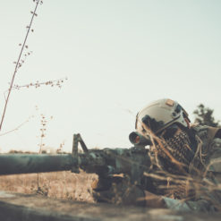 A soldier in camouflage gear lying on the ground with a rifle. The soldier is wearing camouflage gear, including a helmet with a camouflage cover and a face mask. The rifle is resting on a bipod and has a scope attached to it. The background is a field with dry grass and a barbed wire fence. The image has a vintage filter applied to it, giving it a faded and aged look. The image is taken from a low angle, looking up at the soldier.