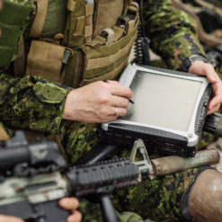 A person in military uniform wearing a green camouflage uniform with a bulletproof vest and rifle sits on the ground with legs crossed. They hold a black, ruggedized tablet device in their hands. The background is a sandy terrain with rocks and shrubs.