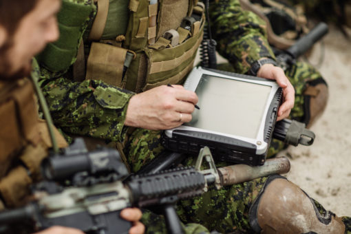 A person in military uniform wearing a green camouflage uniform with a bulletproof vest and rifle sits on the ground with legs crossed. They hold a black, ruggedized tablet device in their hands. The background is a sandy terrain with rocks and shrubs.