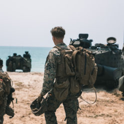 Two soldiers in camouflage uniforms and large backpacks are patrolling a beach. In the background, armored military vehicles with antennas are visible, suggesting a temporary base or staging area. The ocean extends into the horizon, adding to the desolate yet serene atmosphere. The photo’s desaturated color palette enhances the solemnity of the scene.