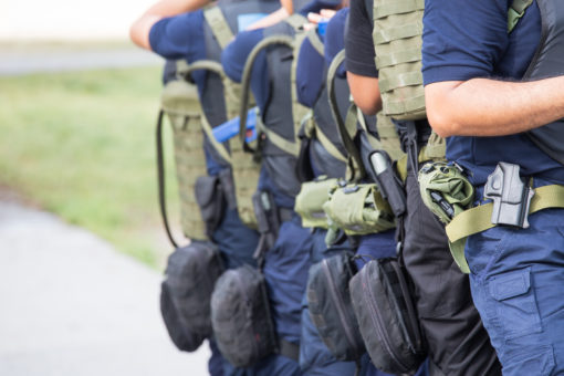Image of military personnel in uniform with equipment, including backpacks and helmets, standing in a line on a sidewalk. The personnel are wearing blue uniforms with black boots and green backpacks. The personnel are also wearing helmets with straps and goggles. The background is a sidewalk with grass on one side and a road on the other. The image is taken from the side and the personnel are facing away from the camera.