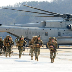 Soldiers wearing camouflage uniforms and carrying backpacks and rifles walking in front of CH-53E Super Stallion helicopter with ‘MARINES’ written on side on airfield with clear sky background