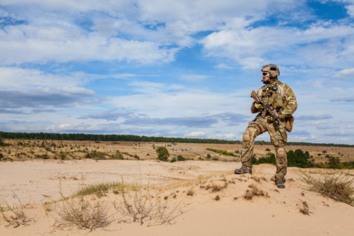 A soldier in full gear is seen walking on a sandy desert. The soldier, clad in a camouflage uniform and helmet, carries a rifle. The desert is dotted with shrubs and bushes, under a blue sky with white clouds. The image conveys a serious and tense mood, indicative of the soldier’s mission.