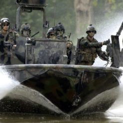 This image depicts a high-speed military boat in action on a river or lake. The boat, camouflaged in a grey and black pattern, is creating a large wake behind it due to its speed. On board, soldiers equipped with helmets and weapons are visible, ready for their mission. The background reveals the natural environment of the operation with trees lining the shore.