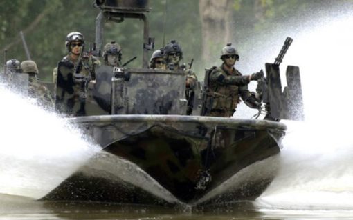This image depicts a high-speed military boat in action on a river or lake. The boat, camouflaged in a grey and black pattern, is creating a large wake behind it due to its speed. On board, soldiers equipped with helmets and weapons are visible, ready for their mission. The background reveals the natural environment of the operation with trees lining the shore.