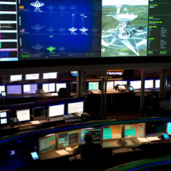 This image depicts an advanced control room, bathed in a soft glow of blue and green lights. The room is filled with multiple workstations, each equipped with several computer monitors displaying a variety of data, graphs, and 3D models. The desks and chairs are sleek and modern, colored in black. Dominating the room is a large screen at the front, showcasing a detailed 3D model of a drilling rig, indicating a high level of technical operation.