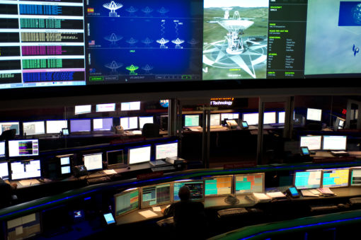 This image depicts an advanced control room, bathed in a soft glow of blue and green lights. The room is filled with multiple workstations, each equipped with several computer monitors displaying a variety of data, graphs, and 3D models. The desks and chairs are sleek and modern, colored in black. Dominating the room is a large screen at the front, showcasing a detailed 3D model of a drilling rig, indicating a high level of technical operation.