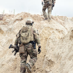 This black and white image captures two soldiers in full gear, advancing down a sandy hill. The soldier in the foreground, identifiable by his camouflage uniform and helmet, is carrying a rifle. The background reveals the challenging terrain they navigate, characterized by sandy hills dotted with rocks and shrubs.