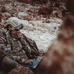 A person in a United States military camouflage uniform and helmet is seated on a rocky terrain, resting against a large rock. The helmet features a protective visor, symbolizing readiness and vigilance. The individual is seated on a blue mat, providing a stark contrast to the surrounding environment. In the blurred background, another figure can be discerned, adding a sense of depth and scale to the scene.