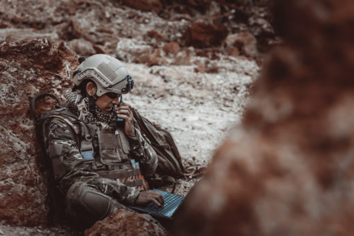 A person in a United States military camouflage uniform and helmet is seated on a rocky terrain, resting against a large rock. The helmet features a protective visor, symbolizing readiness and vigilance. The individual is seated on a blue mat, providing a stark contrast to the surrounding environment. In the blurred background, another figure can be discerned, adding a sense of depth and scale to the scene.