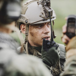 A U.S. military personnel is depicted in full field gear, standing amidst a verdant outdoor setting. The individual is clad in a camouflage jacket and a helmet equipped with a mount for potential camera or night vision goggles usage. A microphone is attached to the helmet, suggesting communication capabilities. The person’s right hand holds a device, possibly a GPS or walkie-talkie, indicating navigational or communication tasks.