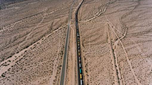 This is an aerial view of a train traveling through a barren desert with little vegetation. The train is on a single track and is surrounded by sand and shrubbery. The train is made up of multiple cars of different colors and is traveling in a straight line.