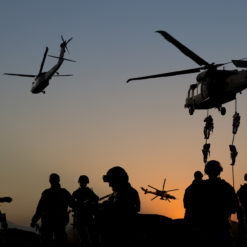 A dramatic scene captured at sunset, displaying four military helicopters in varying stages of flight against a dusky sky. Two helicopters are prominently visible: one is hovering closer to the ground with two sets of soldiers fast-roping down, their silhouettes stark against the dimming light, while another helicopter is ascending, slightly tilted. In the background, two more helicopters can be faintly seen, appearing smaller due to distance. On the ground, several soldiers' silhouettes are visible, some standing still, others in movement, all set against the gradient of the sunset horizon.