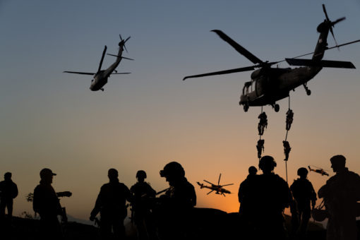 A dramatic scene captured at sunset, displaying four military helicopters in varying stages of flight against a dusky sky. Two helicopters are prominently visible: one is hovering closer to the ground with two sets of soldiers fast-roping down, their silhouettes stark against the dimming light, while another helicopter is ascending, slightly tilted. In the background, two more helicopters can be faintly seen, appearing smaller due to distance. On the ground, several soldiers' silhouettes are visible, some standing still, others in movement, all set against the gradient of the sunset horizon.