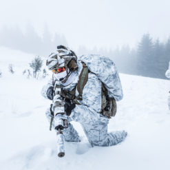 Two United States military personnel in winter camouflage, one kneeling and one standing, during a training exercise in a snowy landscape. The personnel are wearing white and gray camouflage clothing and carrying weapons. The background is a snowy landscape with trees and hills in a foggy and overcast setting.