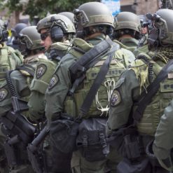 A group of police officers in riot gear, with helmets, shields, and weapons, standing in a line on a city street. The background consists of buildings and trees.