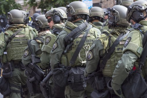 A group of police officers in riot gear, with helmets, shields, and weapons, standing in a line on a city street. The background consists of buildings and trees.