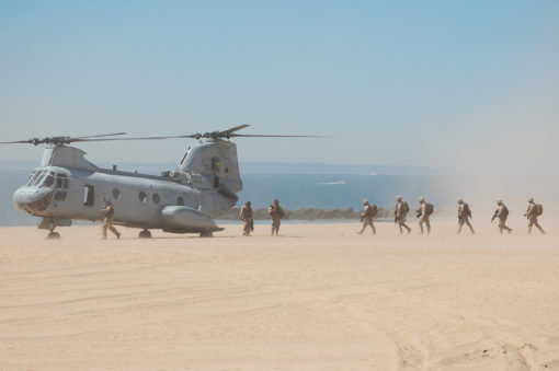 A group of United States military personnel are seen disembarking from a CH-53E Super Stallion helicopter on a sandy beach. The helicopter is gray in color and has two rotors. The personnel are wearing camouflage uniforms and carrying backpacks. The background is a sandy beach with the ocean visible in the distance. The sky is clear and blue.