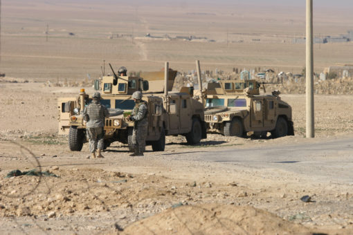 Two tan military vehicles are parked on a dirt road with personnel standing nearby. The vehicles appear to be armored and have turrets on top. The background consists of a flat desert landscape with power lines and a road in the distance.