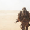 Image of a United States military personnel walking in a desert environment with full gear and equipment. The personnel is wearing a helmet, backpack, and a rifle. The background consists of a sandy desert with a clear sky. The image has a faded and hazy effect, likely due to the desert environment.